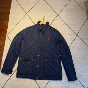 Mörkblå quiltad jacka från U.S. Polo ASSN. - Snygg mörkblå quiltad jacka från polo med rött broderat logomärke på bröstet. Jackan har två stora fickor med knappar, ståkrage och knappstängning framtill. 