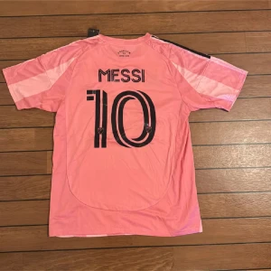 Inter Miami Messi #10 rosa fotbollströja - Säljer en officiell Inter Miami fotbollströja från Adidas i slim fit, storlek M. Tröjan är rosa med svarta detaljer, har korta ärmar och randigt mönster. Messi #10 tryck på ryggen, klubbmärke och Adidas-logga på bröstet. Tillverkad i lätt polyester.