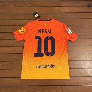 Barcelona Messi #10 orange matchtröja - Säljer en officiell FC Barcelona matchtröja från Nike med Messi och nummer 10 på ryggen. Tröjan är orange med gul toning och har tryck från Qatar Foundation och UNICEF. Kortärmad, med klubbmärke och La Liga-patch på ärmen. Tillverkad i lätt Dri-FIT material.