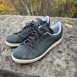 Adidas Stan Smith svarta sneakers - Svarta Adidas Stan Smith sneakers med vit sula och vita detaljer vid hälen. Skorna har klassisk låg profil, rund tå och snörning med svarta och vita mönstrade skosnören. Ovandelen är i skinn och insidan är vit. Perfekt för dig som gillar ikoniska och stilrena sneakers.