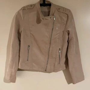 Beige bikerjacka från Divided H&M - Snygg beige bikerjacka från Divided by H&M i storlek 36. Jackan har asymmetrisk dragkedja, axelklaffar och dragkedjor vid fickor och ärmslut. Perfekt för dig som vill ha en stilren och trendig look.