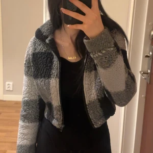 Grå och svart teddyjacka från SHEIN - Säljer en mysig teddyjacka från SHEIN i storlek XS. Jackan har ett rutigt mönster i olika nyanser av grått och svart, croppad passform och dragkedja framtill. Perfekt för dig som gillar chill och trendiga vibes. Hög krage och långärmad modell.