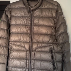 Moncler Acorus  - Snygg mörkgrå dunjacka från Moncler med quiltad design och klassisk logga på ärmen. Jackan har dragkedja framtill och två sidofickor med dragkedja. Perfekt för kalla dagar och har en stilren look som funkar till alla dagar. Negativt är att det finns små fläckar på ryggen som knappast syns. Storlek 4/L och såklart äkta. 