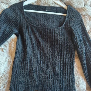 Svart ribbad långärmad topp Gina Tricot - Svart långärmad topp från Gina Tricot med ribbad struktur och fyrkantig halsringning. Materialet är mjukt och stretchigt, vilket gör den bekväm och följsam. Perfekt att styla med jeans eller kjol för en cool och enkel look.