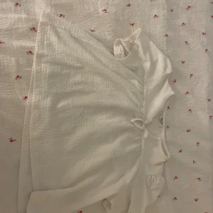 Vit blus med knyt från Kappahl - Supersöt vit blus från Kappahl i strukturerad bomull. Modellen har kort puffärm och en fin knytning framtill vid halsringningen. Perfekt för en fräsch och enkel look med romantiska vibbar.