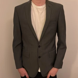 Grå kavaj från J.Lindeberg - Snygg grå kavaj från J.Lindeberg med klassisk passform och två knappar framtill. Kavajen har två fickor med lock och en bröstficka. Materialet är mjukt och har en lätt struktur, perfekt för en stilren look. Fodret är slätt och mörkt, vilket ger en exklusiv känsla. ALDRIG ANVÄND och PRIS ÄR DISKUTERBART nypris:4500