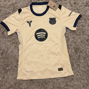 Beige FC Barcelona Nike fotbollströja small spelar version  -  Spelar version Snygg beige FC Barcelona fotbollströja från Nike med svarta och blå detaljer på krage och ärmslut. Tröjan har korta ärmar, Dri-FIT ADV-material och tryck med Spotify, UNHCR och klubbmärke. Perfekt för dig som vill ha en unik Barca-tröja med modern design.