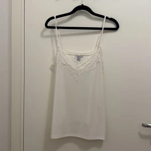 Vit linnetopp med spets från Amisu/new Yorker - Säljer en vit linnetopp från Amisu med tunna axelband och v-ringning dekorerad med vacker spets. Toppen har en enkel och stilren design som passar perfekt till jeans eller kjol. Materialet känns mjukt och lätt mot huden.