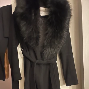 Svart kappa med päls från Chiara Forthi - Snygg svart kappa från Chiara Forthi Milano med stor fluffig pälsimitation vid kragen, silverfärgade knappar och bälte i midjan. Stilren och elegant design, perfekt för dig som vill ha en statement piece. Långärmad och med klassisk passform.