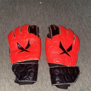 Röda målvaktshandskar GK200 strl 6 - Säljer ett par röda och svarta målvaktshandskar från GK200 i storlek 6. Handskarna har hexagonmönster på ovansidan, svart logga och breda kardborreband vid handleden för extra stöd. Perfekta för fotbollsmålvakter som vill sticka ut på planen.