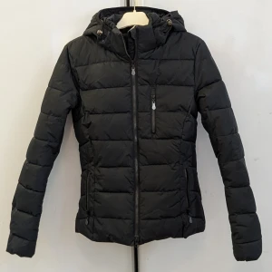RIDJACKA , JH Collection XS - ANVÄND EN ENDA GÅNG. Svart pufferjacka från JH Collection i storlek XS. Jackan har huva med snörning, flera dragkedjeförsedda fickor och quiltad design. Perfekt för kalla dagar med sin stilrena look och figurnära passform. Snygg vinterridjacka 
