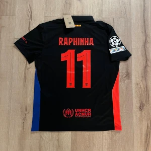 FC Barcelona Raphinha matchtröja Nike - Snygg FC Barcelona fotbollströja med Raphinha och nummer 11 på ryggen. Svart bas med röda och blå detaljer, officiella loggor från Nike, Spotify och UNHCR. Kortärmad modell i lätt och ventilerande material, perfekt för match eller träning.
