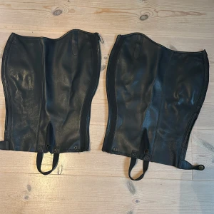 Svarta skinnchaps storlek L - Säljer ett par svarta chaps i skinn, perfekta för ridning. De har dragkedja längs sidan och elastiska remmar vid foten för att sitta stabilt över ridstövlar. Insidan är mjuk och mockaliknande för extra komfort. Passar dig som vill ha en snygg och funktionell look i stallet.