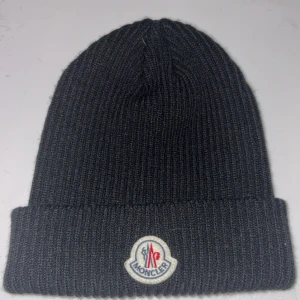 Svart Moncler mössa - Svart ribbstickad mössa från Moncler med uppvikt kant och klassisk Moncler-logga framtill. Mössan är tillverkad i mjukt material och har en enkel, stilren design som passar perfekt för kalla dagar.