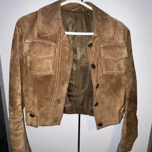 Mockajacka från Jofama, brun - 70tals mocksjacka, snyggt croppad mockajacka från Jofama i brun färg. Jackan har stora fickor med lock framtill, markerad krage och knäppning med mörka knappar. Materialet är mjuk mocka och insidan är fodrad med slätt tyg. Perfekt för dig som gillar retrovibe och klassisk stil. Det står storlek 40 på insidan, men eftersom att jackan är gammal kan det inte vara exakt som en 40 jacka nu för tiden. 