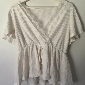 Vit omlottblus med spets från SHEIN - Supersöt vit omlottblus från SHEIN med korta ärmar, knyt i midjan och volang nertill. Blusen har spetsdetaljer längs v-ringningen och är gjord i ett strukturerat bomullstyg. Perfekt för en fräsch och romantisk look.