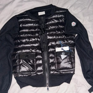 Svart quiltad cardigan jacka Moncler - Svart cardigan jacka från Moncler med quiltad, glansig front och stickade ärmar. Jackan har två fickor med knappstängning, rund hals och dragkedja framtill. Moncler-logga på ärmen och snygga detaljer som ger en clean och modern look. Skriv gärna om ni verkar intresserade 🫰🤗. Postar samma inom en till två dagar efter ni köpt💕. 
