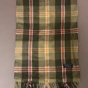 Rutig halsduk från Burberry - Snygg rutig halsduk från Burberry i klassiska gröna, beige och röda toner med inslag av vitt och gult. Halsduken har fransar i ändarna och är tillverkad i mjuk ull, perfekt för kyliga dagar. Den ikoniska Burberrys-loggan sitter på en liten etikett. halsduken skickad från Norge och moms kan tilkomma. betalning måste ske via paypal eller liknande