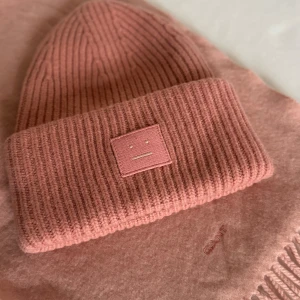 Rosa ribbstickad mössa Acne Studios - Rosa ribbstickad mössa från Acne Studios på Norrmalmstorg, med deras ikoniska face-patch framtill. Tillverkad i mjuk ullblandning. Matchande halsduk från Gant följer med (2 mindre hål i, se bild).
