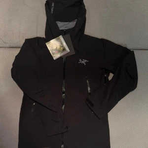 Svart Arc'teryx jacka  - Arc'teryx svart vindjacka i herrmodell med Gore-Tex Paclite Plus-material. Jackan har huva, vattentäta dragkedjor och två sidofickor med zip. Snygg logga på bröstet och Arc'teryx-tryck på huvan. Perfekt för regniga dagar och outdoor.