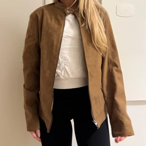 Brun mockajacka från  - Snygg brun mockajacka med stilren design. Jackan har ståkrage med knapp, dragkedja framtill och två sidofickor. Materialet är mjuk mocka och passformen är normal. Perfekt för dig som gillar en clean och klassisk look.