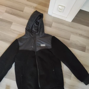 Svart teddyjacka med huva från Smalldook - Svart teddyjacka från Smalldook med huva och snygga detaljer i blankt syntetmaterial över axlar och bröst. Jackan har hel dragkedja framtill, ribbade muddar och en chill, loose passform. Perfekt för kalla dagar och streetwear-stil.