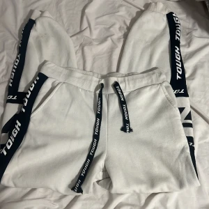 Vita mjukisbyxor  - Vita mjukisbyxor med breda svarta revärer längs sidorna där det står 'TOUGH' i vitt. Byxorna har resår i midjan och snörning med matchande svart band. Mjukt material och avslappnad passform, perfekta för chill dagar. Små fläckar längst ner på benen men syns knappt