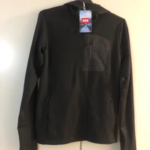 Ny svart Helly Hansen Versalite Hooded Jacket med etikett - Helt ny svart Helly Hansen Versalite Hooded Jacket med etikett säljes.. Svart hoodie från Helly Hansen med huva och dragkedja framtill. Tröjan har en bröstficka med dragkedja och två sidofickor. Regular fit och långärmad, perfekt för chill eller outdoor. Materialet är mjukt och känns som polyester. Snygg och enkel design med HH-logga på bröstet.