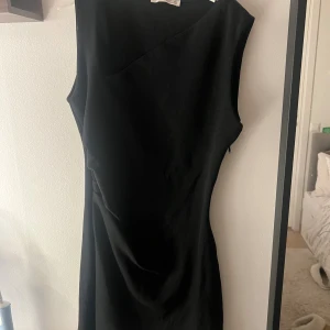 Svart ärmlös klänning med asymmetrisk hals - Snygg svart klänning med asymmetrisk halsringning och figurnära passform. Klänningen är ärmlös och har en stilren, minimalistisk design som ger en cool och modern vibe. Perfekt för dig som gillar enkla men trendiga plagg.