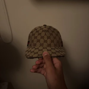 Gucci keps med GG-mönster - Snygg keps från Gucci i beige med klassiskt GG-monogram över hela kepsen och en grön-röd rand på sidan. Justerbar rem baktill och insida med svart tyg. Perfekt för dig som vill sticka ut med en lyxig accessoar. Inga deffekter med kepsen, den har fortfarande bra form. Dustbag finns.