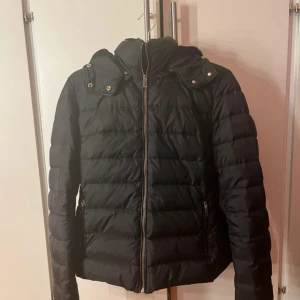 Svart pufferjacka med huva - Svart pufferjacka med avtagbar huva och dragkedja framtill. Jackan har quiltad design och två sidofickor med dragkedja. Perfekt för kyliga dagar och har en stilren look som funkar till många outfits. Storlek XL men skulle mer säga att den passar M/L då den är liten i storleken