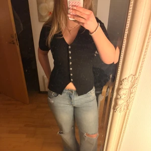 Svart v-ringad topp från Gina Tricot - Svart kortärmad topp från Gina Tricot med v-ringning och snygga knappar framtill. Modellen är figurnära med lätt utsvängd nederkant och en liten slits framtill. Materialet är mjukt och stretchigt som stickat, perfekt för en chill och stilren look. Använd fåtal gånger!💞