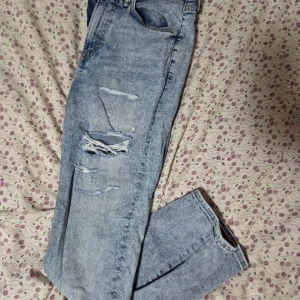 Skinny croppade jeans från &Denim - Snygga ljusblå skinny jeans från &Denim i croppad modell. De har slitna detaljer och hål på benen för en cool, trendig look. Klassisk femficksdesign, dragkedja och knapp framtill. Perfekta för dig som gillar streetstyle.