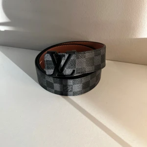 skärp svart/grå -  bälte i klassiskt Damier-mönster i svart och grått. Snygg logga i svart metall på spännet. Bältet är i exklusivt med brun insida och tydliga sömmar. Perfekt accessoar för att lyfta din outfit.