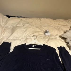 Mörkblå stickad tröja från Lacoste - Säljer en mörkblå stickad tröja från Lacoste med klassisk rund halsringning och diskret krokodillogga på bröstet. Tröjan har lång ärm och är ribbstickad för en snygg och enkel look. Perfekt för dig som gillar stilrena plagg med premiumkänsla.