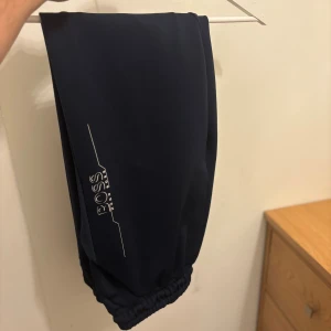 Mörkblå mjukisbyxor från Hugo Boss - Snygga mörkblå mjukisbyxor från Hugo Boss med vit logga längs benet. Byxorna har elastisk mudd vid benslut och är gjorda i mjukt syntetmaterial. Perfekta för chill eller träning, med en loose passform och stilren design.