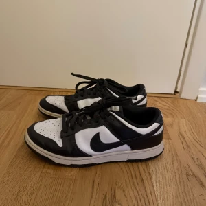 Nike Dunk Low svart/vit sneakers - Nike Dunk Low sneakers i svart och vitt med klassisk låg siluett. Skorna har perforerad tå, snörning och den ikoniska swoosh-loggan på sidan. Tillverkade i skinn med platt sula och rund tå. Perfekt för dig som gillar streetwear och sportig stil.