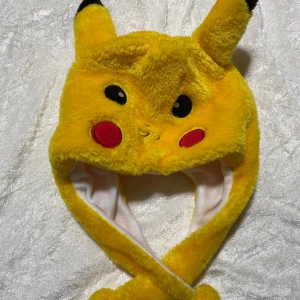 Gul Pikachu mössa med öron - Supermysig gul Pikachu mössa med stora öron och broderade ansiktsdetaljer. Mössan är gjord i mjuk syntetpäls och har långa flikar som kan korsas under hakan. Perfekt för dig som älskar Pokémon och vill sticka ut!  