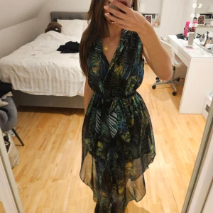 Bladmönstrad långklänning med knyt - Snygg långklänning med tropiskt bladmönster i grönt, blått och gult. Klänningen är ärmlös, har v-ringning och markerad midja med knytband. Tunt, luftigt material som ger en flowy känsla. Perfekt för sommarens varma dagar.