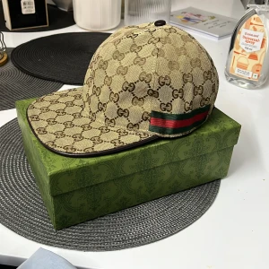 Gucci keps med GG-mönster och röd/grön detalj - Exklusiv keps från Gucci i beige och brunt med klassiskt GG-monogram över hela kepsen. Kepsen har en böjd skärm, bruna läderdetaljer och den ikoniska röd-gröna randen på sidan. Tillverkad i bomullscanvas med insida i brunt tyg och justerbar rem bak.