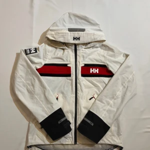 Vit vindjacka från Helly Hansen - Snygg vit vindjacka från Helly Hansen med huva och svarta detaljer på ärmsluten. Jackan har röda och svarta ränder över bröstet, HH-logga på fram- och baksidan samt dragkedja framtill. Perfekt för dig som gillar sportig stil och funktionella plagg.