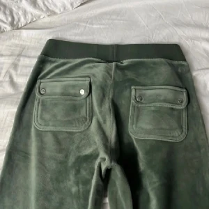 Gröna mjukisbyxor i velour med snörning - Mörkgröna mjukisbyxor i mjuk velour med bred resår i midjan och snörning framtill. Byxorna har stora fickor både fram och bak, bakfickorna har lock och knappar. Liten broderad logga framtill på höger sida.