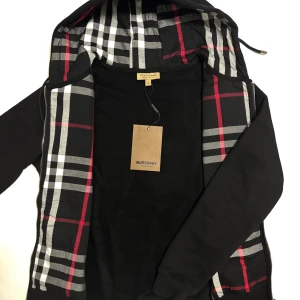 Svart Burberry kofta  - Svart kofta från Burberry 10/10 kvalité med klassiskt rutig insida i svart, vitt och rött. Koftan har huva med dragsko, dragkedja framtill och diskret broderad logga på bröstet. Perfekt för de kommande kyliga dagar. Storleken är M. Skickas inom 24timmar! 