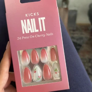 KICKS Nail It Press On Cherry Nails - 24-pack press on-naglar i mandelform med rosa och vit ombre samt söta körsbärs- och blomdetaljer. Perfekt för dig som vill ha snygga naglar snabbt utan att behöva måla själv. Lätta att applicera och ta bort.