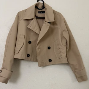 Beige croppad skepparkavaj från Zara - Snygg beige croppad skepparkavaj från Zara med dubbelknäppning och klassisk krage. Jackan har svarta knappar, två fickor fram och långa ärmar med detalj vid mudden. Perfekt för dig som gillar stilrena och trendiga plagg.