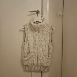 Beige pälsväst från Amisu med dragkedja - Mysig beige pälsväst från Amisu med hög krage och dragkedja framtill. Västen har ribbade detaljer vid krage, ärmöppningar och nederkant samt två fickor fram. Insidan är fodrad i satin för extra komfort. Perfekt för lager-på-lager och chill stil.