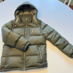 Grön pufferjacka från Polo Ralph Lauren - Snygg mörkgrön pufferjacka från Polo Ralph Lauren med huva och orange logga på bröstet. Jackan har tre dragkedjeförsedda fickor fram och quiltad design. Perfekt för kalla dagar och har en modern, normal passform.