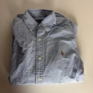 Blå skjorta från Ralph Lauren XS - Snygg ljusblå skjorta från Ralph Lauren i slim fit med klassisk krage och broderad logga på bröstet. Skjortan har långa ärmar och vita knappar framtill. Tillverkad i mjuk bomull som känns skön mot huden. Perfekt för dig som gillar stilrena och tidlösa plagg.