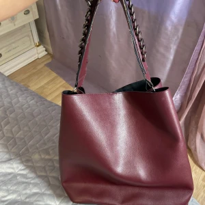 Vinröd axelväska med kedjedetaljer,Oanvänd  - Oanvänd helt ny äkta läder. Jätte fin elegant.Stilren vinröd axelväska i syntetmaterial med breda axelband dekorerade med kedjor. Väskan har ett rymligt huvudfack, innerficka med knappstängning och magnetlås upptill. Perfekt för dig som vill ha plats för allt du behöver i vardagen.