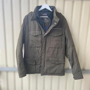 Olivgrön FieldJacket från Snoot - Parkajacka från Snoot i olivgrön färg med flera praktiska fickor, både med lock och dragkedja. Jackan har hög krage, innerfoder i mörkblått och detaljer som metallring och dubbla dragkedjor. Perfekt för dig som gillar funktionell och snygg stil.